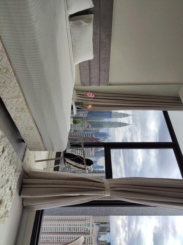 A spacious bedroom is furnished with a large bed and minimalistic decor. Floor-to-ceiling windows provide a view of the KLCC Twin Towers and city skyline. A small seating area with white chairs is near the window, complemented by a vase of flowers on the small table.
