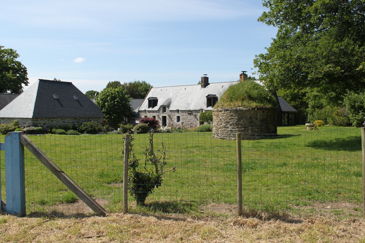 A charming rural property is displayed, featuring a traditional stone building with a sloped roof and multiple windows. A round stone structure is visible nearby, highlighting the unique architecture. Lush greenery surrounds the area, with a wooden fence marking the boundary of the well-kept grounds.