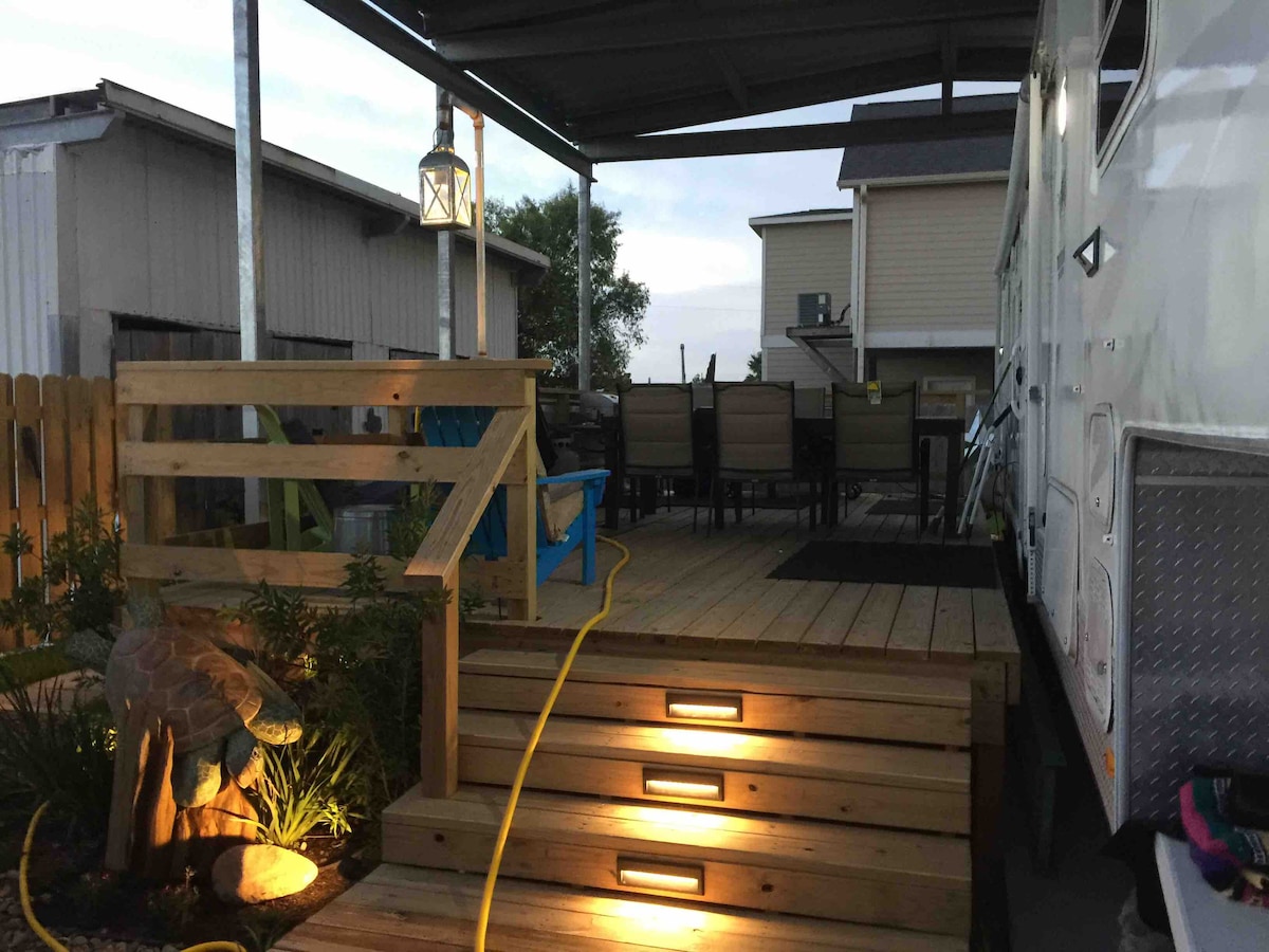 The deck area is shown in the evening light, with wooden steps leading up to the RV. A pathway is illuminated by warm lighting, highlighting the landscaped area. A dining table with chairs is visible in the background beneath a covered area.