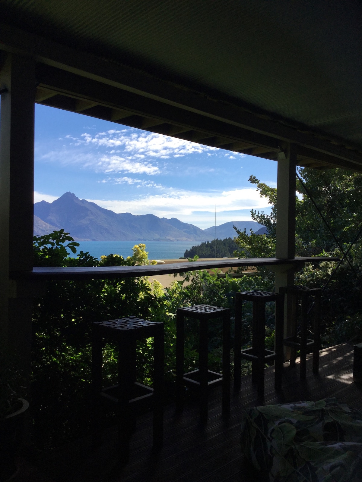 A view is captured from a covered porch, revealing a stunning panorama of Lake Wakatipu and surrounding mountains. The foreground features a set of four wooden stools, framed by lush greenery, creating a tranquil outdoor retreat.