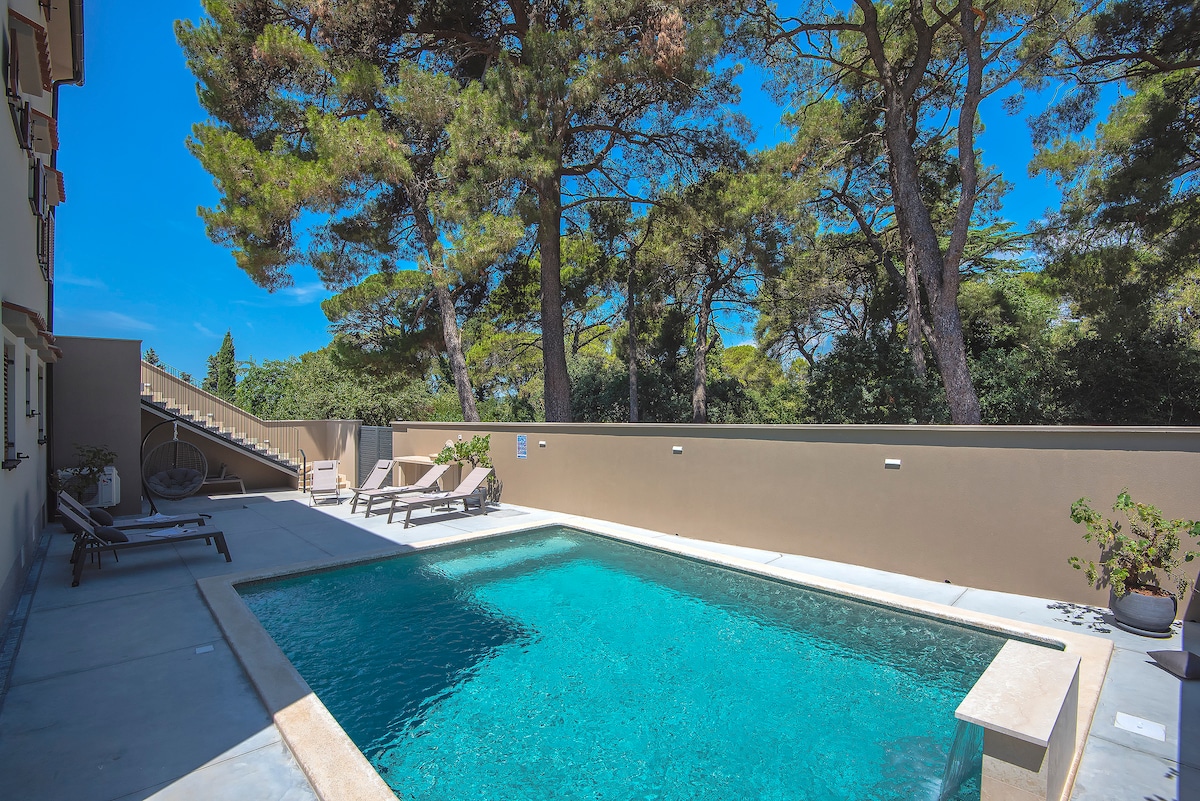 A serene outdoor space features a crystal-clear pool surrounded by a sun terrace. Two sun loungers are positioned nearby, and majestic trees provide shade and natural beauty. The walls are painted in a soft hue, enhancing the tranquil environment.