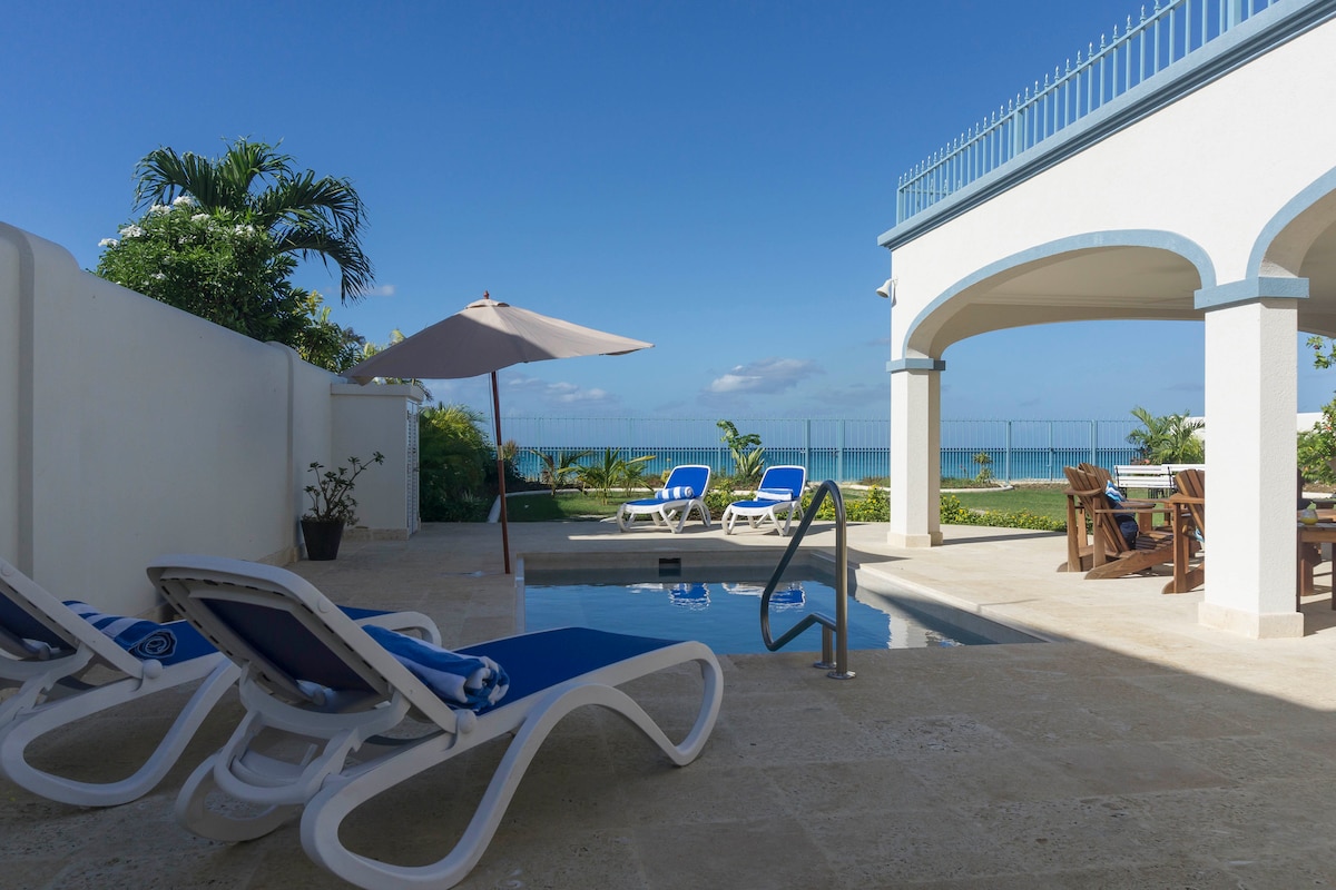 A sleek saltwater pool is framed by lounge chairs, inviting relaxation. An umbrella provides shade near the poolside. Beyond, lush greenery and vibrant ocean views are visible, enhancing the serene outdoor space. A dining area is situated under the archway, connecting the indoor and outdoor environment.