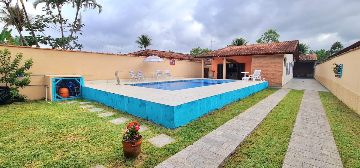 A spacious outdoor area features a well-maintained pool bordered by a blue structure. Several white lounge chairs are positioned poolside, and a shaded area is visible. Green grass and decorative plants enhance the setting, with a pathway leading to a covered porch and outdoor kitchen.