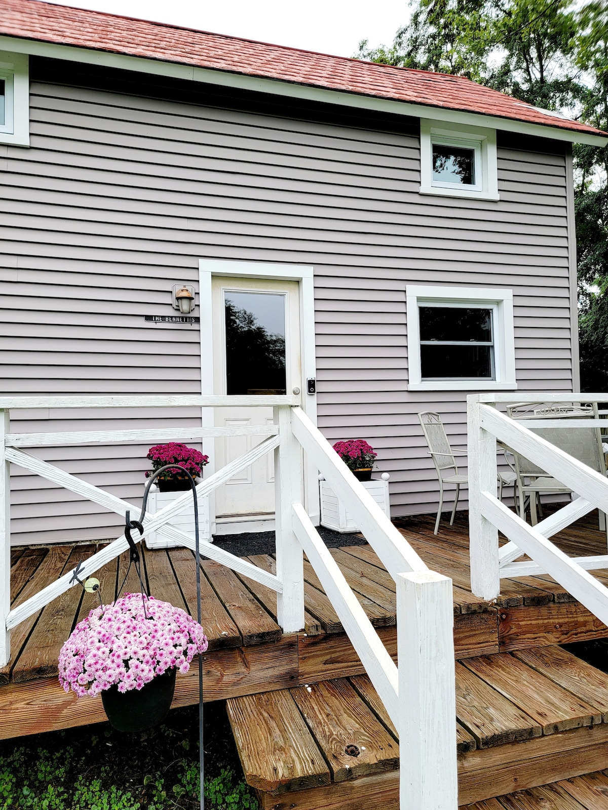 The entrance features a welcoming door framed by two windows, with steps leading up to a wooden porch. Potted flowers in vibrant colors add charm, while a dining set is placed nearby for outdoor enjoyment.