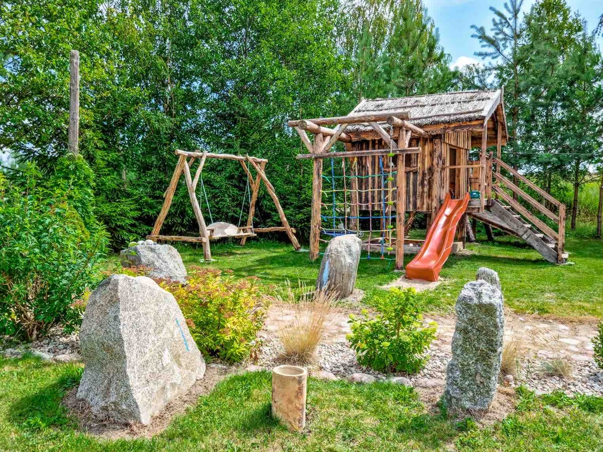 A wooden play structure features a slide, climbing area, and swings, surrounded by a grassy yard. Large rocks and small shrubs are arranged throughout, adding natural elements to the playful setting. Mature trees provide a backdrop, creating a sense of privacy and tranquility.