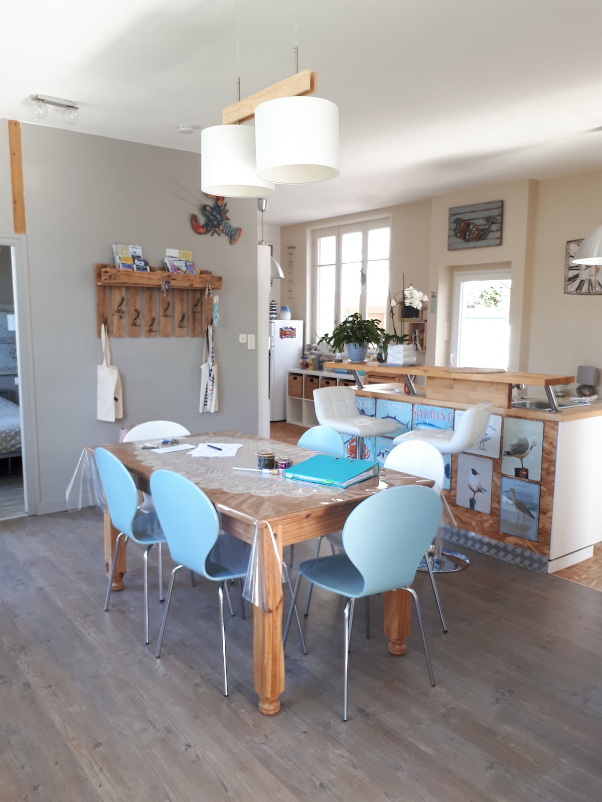 A bright dining area features a wooden table surrounded by six light blue chairs. Natural light pours in through large windows, illuminating the neutral-toned walls. A modern light fixture hangs above the table, while decorative elements are visible on walls and shelves.