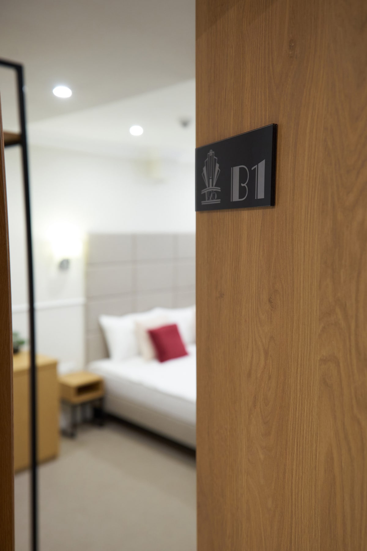 A wooden door is partially open, revealing a modern room with a spacious bed dressed in white linens and adorned with a red accent pillow. Soft lighting and contemporary decor are visible, creating an inviting entrance to the accommodation.