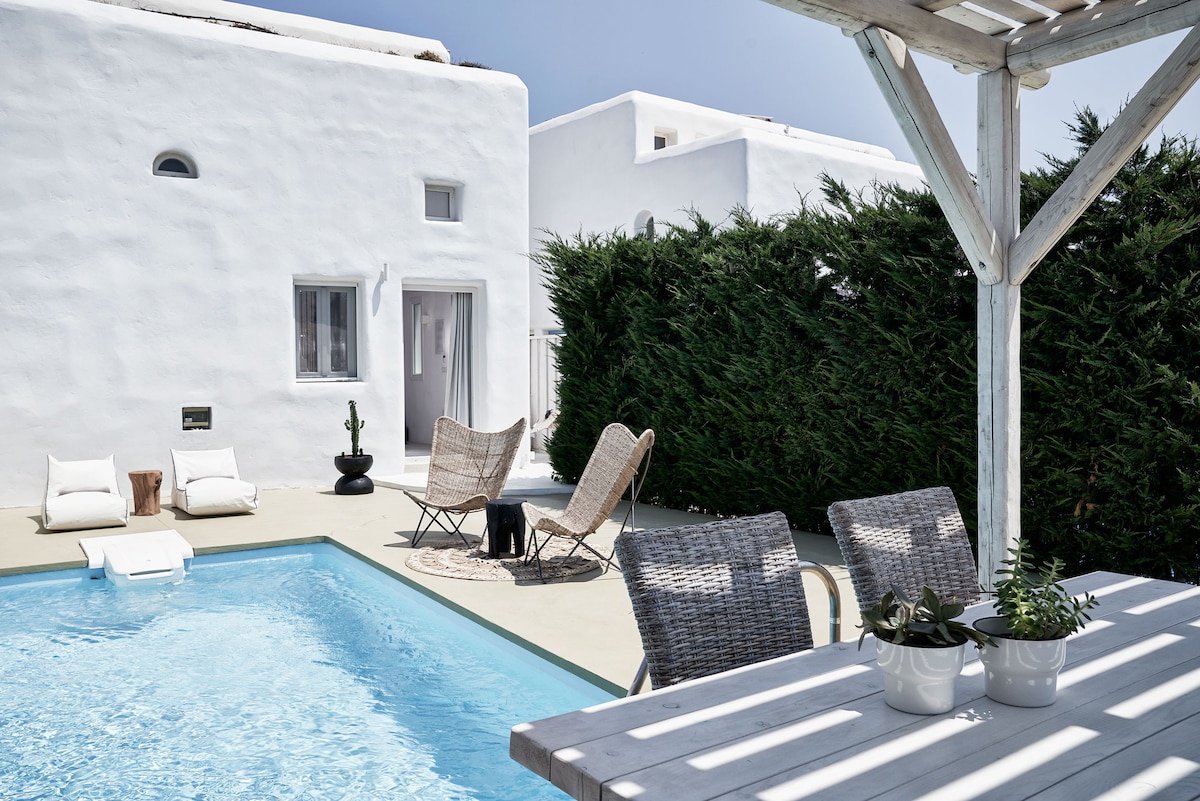 An outdoor area is depicted with a private pool surrounded by light-colored tiles. Two lounge chairs are positioned beside the pool, along with a round rug nearby. A dining table with potted plants is placed under a wooden pergola, complemented by lush greenery in the background.