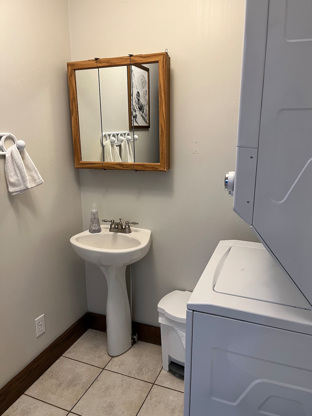 A compact bathroom features a pedestal sink beneath a framed mirror. A stackable washer and dryer are located to the right, with a small wastebasket nearby. White towels hang from a hook, and tiled flooring is present throughout the space.