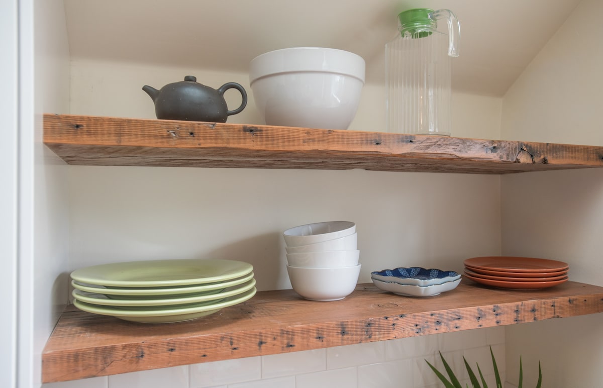 Open shelves are displayed in the kitchen, showcasing various dishware. A black teapot, clear pitcher, and an assortment of bowls in white, green, and blue tones can be seen. The rustic wooden shelves contribute to the cottage's welcoming ambiance.