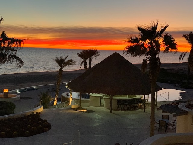 A serene beachfront scene is illuminated by a vibrant sunset, casting warm hues across the sky. Palm trees provide a tropical backdrop to the pool area, where a shaded gazebo with bar seating offers a place to relax. The gentle waves lap at the shore.