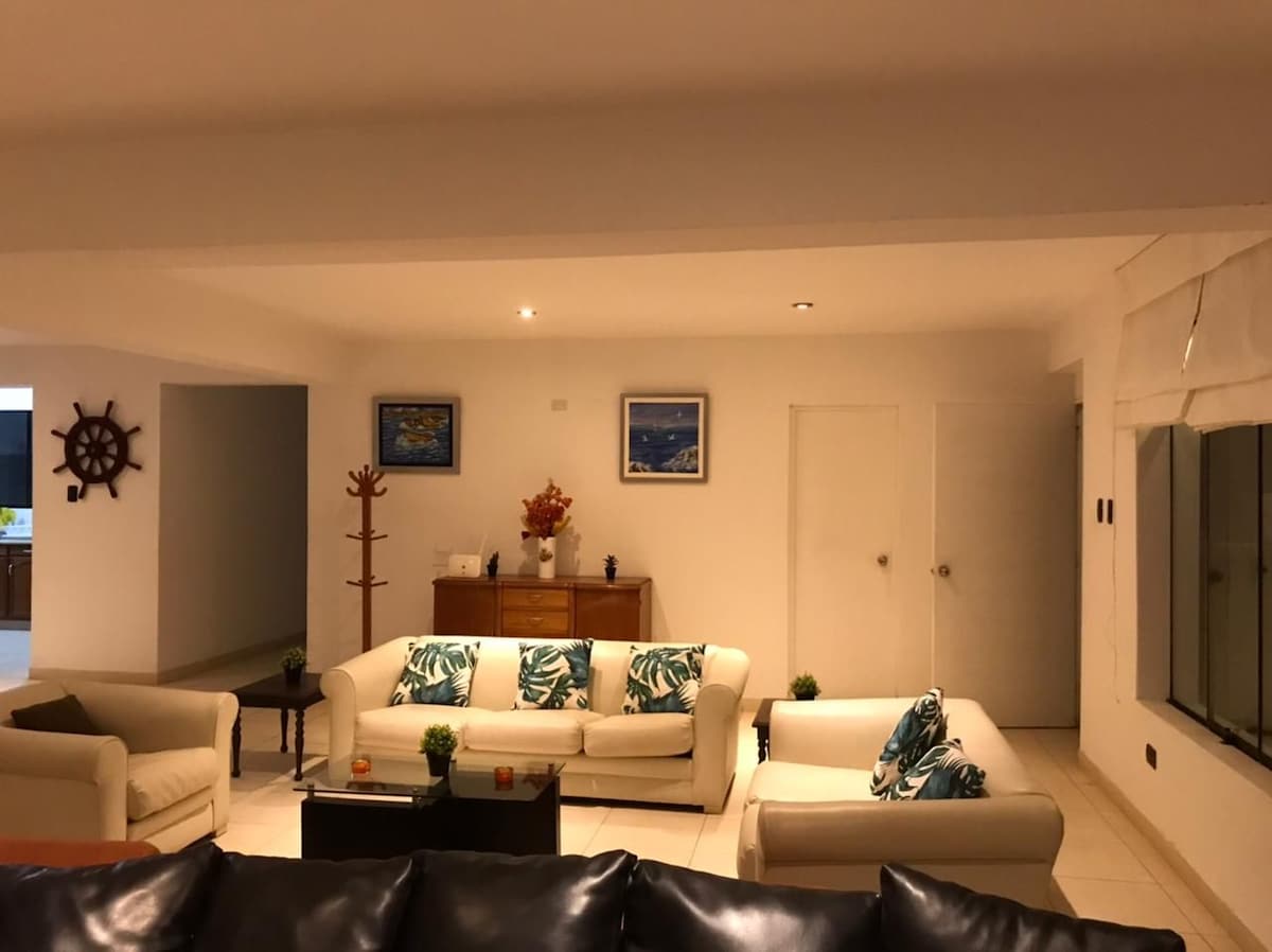 A spacious living area is furnished with light-colored sofas adorned with tropical-patterned cushions. A wooden sideboard is positioned against the wall, complemented by decorative elements. Soft lighting and large windows contribute to a bright and open feel in the room.