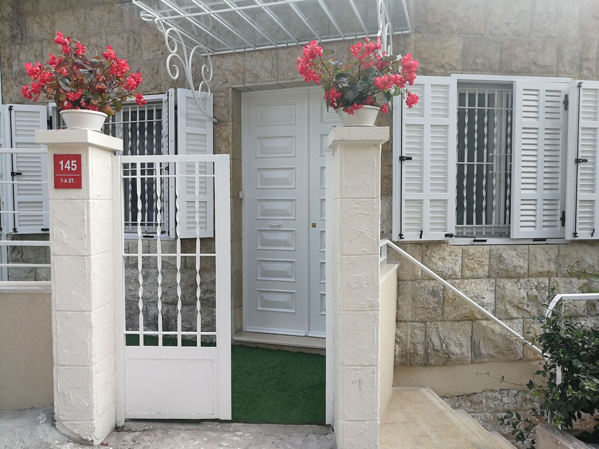 The entrance of a traditional house is framed by white stone walls and adorned with potted red flowers. A white gate leads to a small green area, while closed shutters and a brightly painted door complete the welcoming facade.