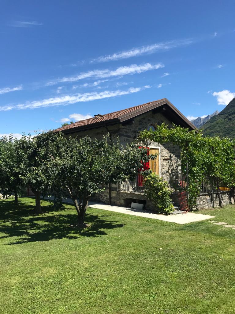 A charming stone cottage is framed by a lush green lawn and trees. A vibrant yellow door adds a pop of color, while the surrounding mountains provide a scenic backdrop under a clear blue sky. The structure exudes a traditional and inviting character.