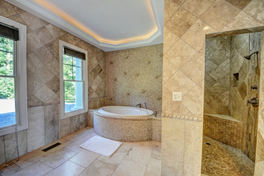 A spacious bathroom features elegant stone tiles and a relaxing jetted tub surrounded by a curved wall. A large shower area is equipped with multiple shower heads. Natural light enters through a well-placed window, complemented by soft, neutral tones throughout the space.