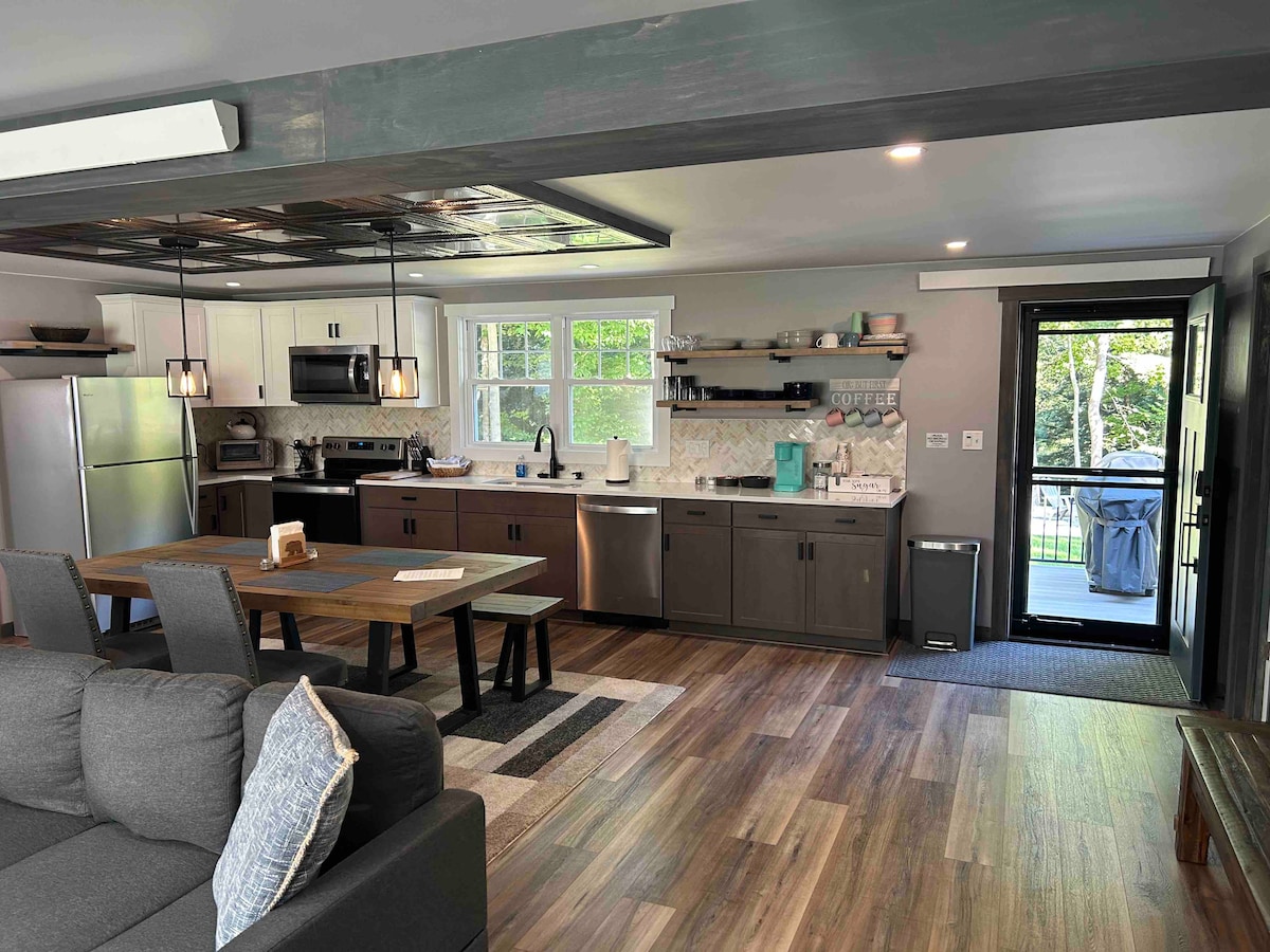 The open-concept kitchen and dining area feature modern appliances and ample counter space. A wooden dining table is accompanied by comfortable chairs. A living area is visible with a cozy couch. Bright light flows in through large windows, connecting the indoor space to the natural surroundings.