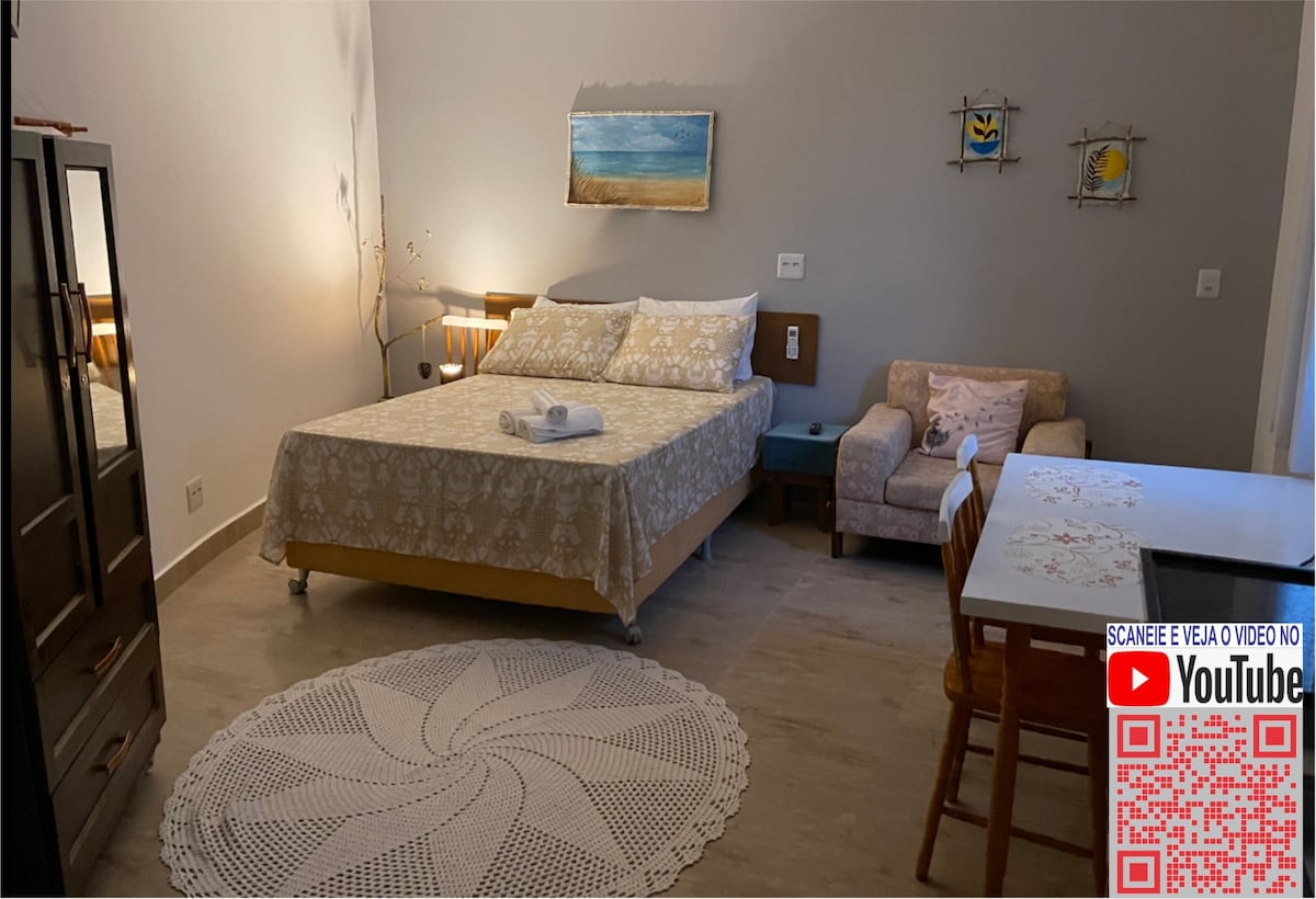 A spacious sleeping area features a large bed covered with a patterned bedspread, accompanied by a small armchair and a wooden table. A handcrafted area rug is centered on the floor, and wall art adds a touch of decor, illuminated by warm lighting.