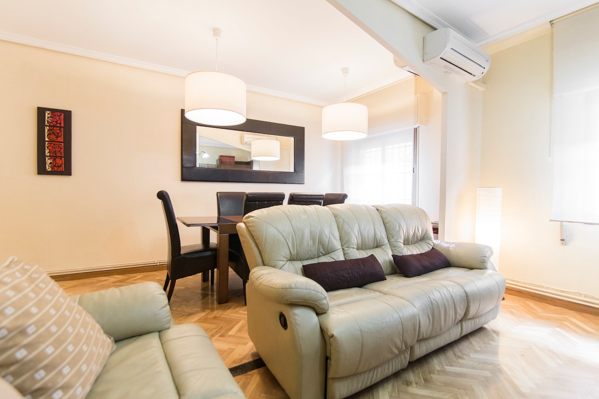 A spacious living area features two comfortable leather sofas paired with dark cushions. A dining table with chairs is positioned nearby under soft lighting from modern fixtures. Large windows allow natural light, enhancing the warm atmosphere of the room.