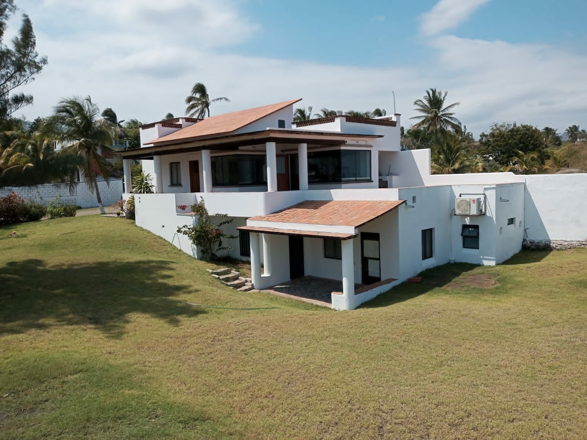 The exterior view of a two-story villa reveals a modern design with a terracotta roof. Large glass windows provide ample natural light. A lush green lawn surrounds the property, and tropical trees are visible in the background, enhancing the serene setting.