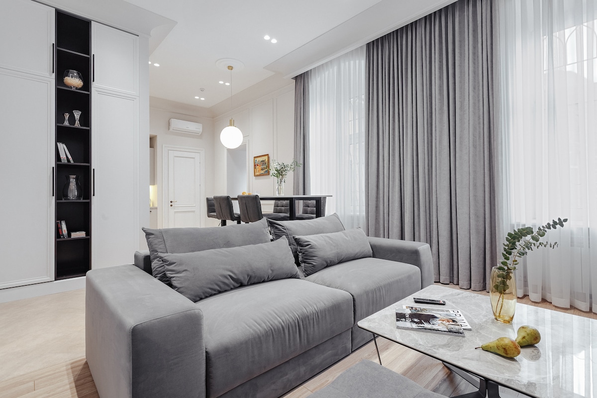 A spacious living area features a comfortable grey sofa, complemented by a marble coffee table topped with decorative items. Large windows are adorned with sheer curtains, allowing natural light to fill the room. A dining area with dark chairs is visible in the background.