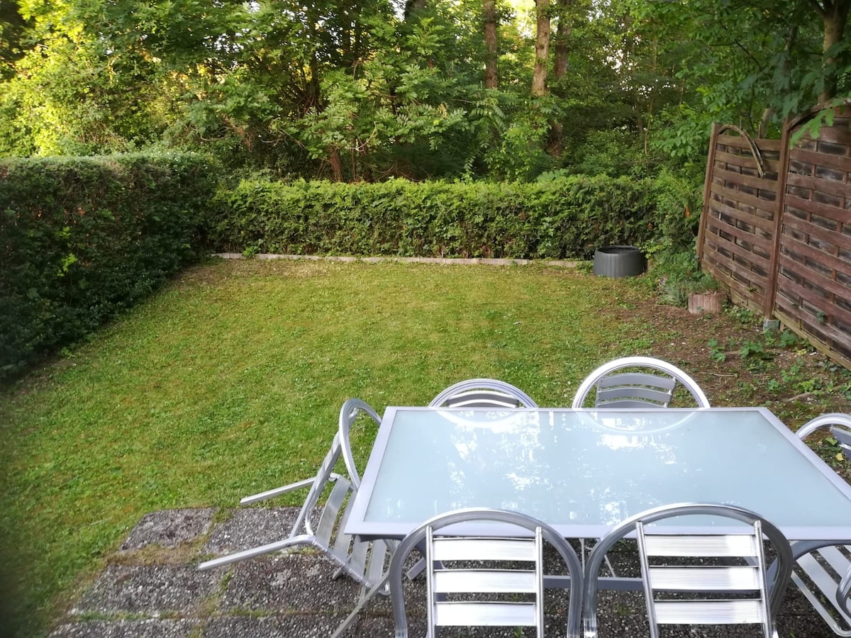 A serene garden area features a well-kept lawn surrounded by lush greenery. A glass-top dining table, accompanied by sleek metal chairs, is positioned on a stone patio. The scene is complemented by a tidy hedge along the perimeter, creating a tranquil outdoor space.