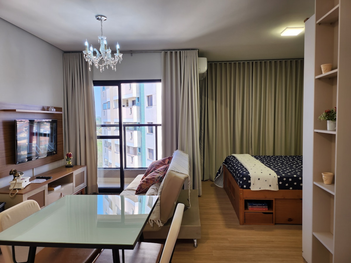 A well-lit studio features a spacious sleeping area with a level bed, flanked by stylish curtains. A light-colored sofa sits opposite a wooden entertainment unit, where a television is displayed. A dining table with chairs is positioned nearby, offering a view through large glass doors.