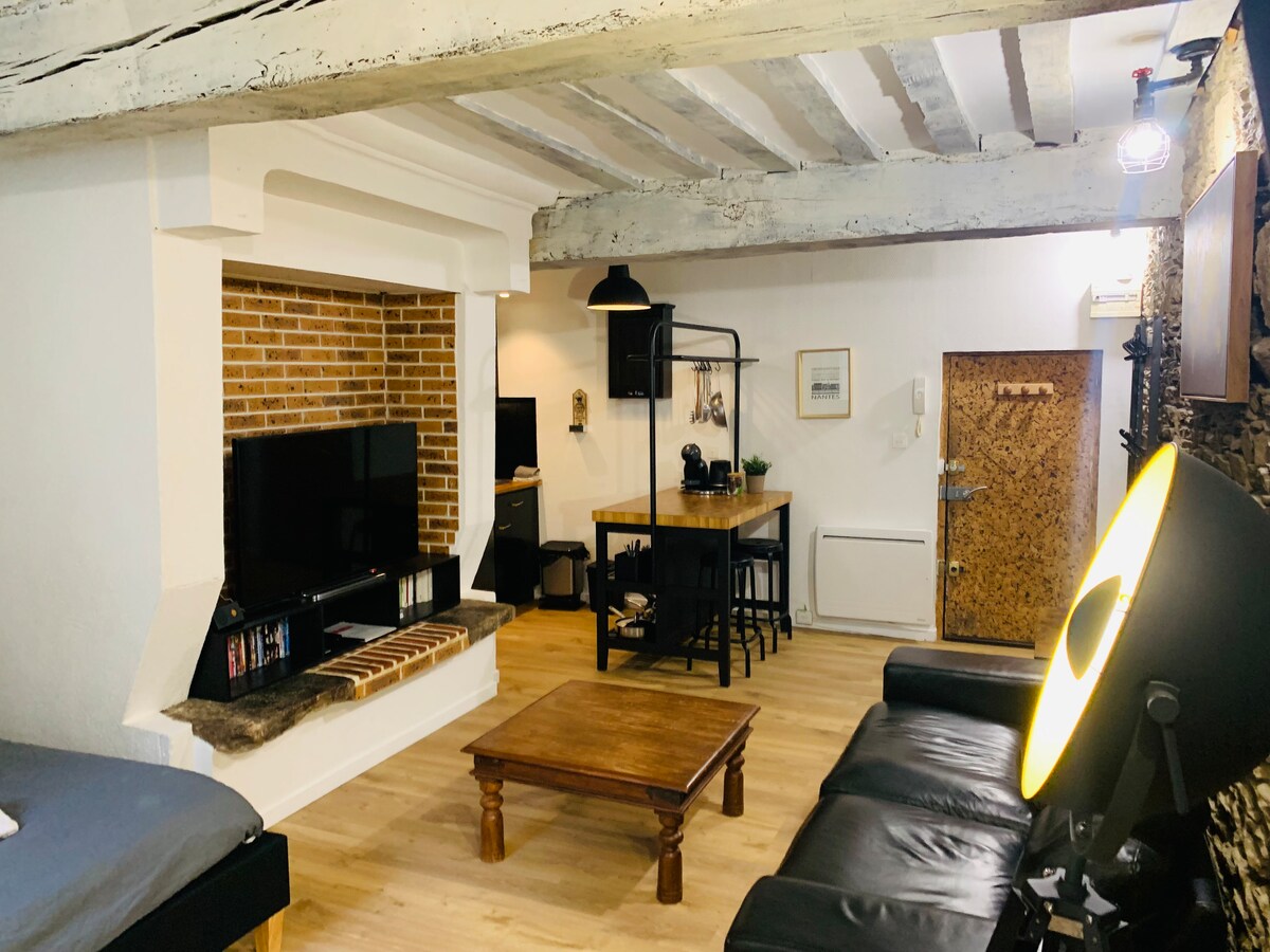 An open living area features a brick fireplace and a connected entertainment unit with a television. A wooden dining table with black stools is visible, alongside a comfortable black sofa. Natural light fills the space through a nearby window, highlighting the wooden floors and exposed beams.