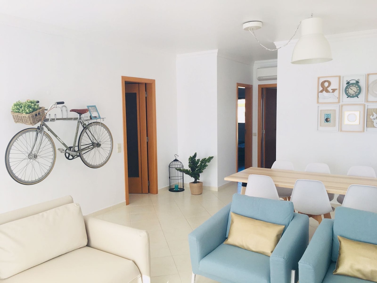A modern living area features light-toned furniture, including two blue chairs and a white sofa. A bicycle is mounted on the wall, and a dining table is set for six. Décor elements include framed art and a decorative birdcage, enhancing the inviting space.