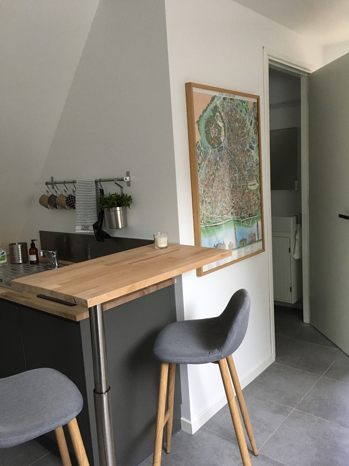 A modern kitchen space is featured, showcasing a wooden countertop with two gray stools positioned nearby. A wall-mounted rack holds various kitchen utensils, while a map is displayed inside a frame. A doorway leads to a nearby bathroom, featuring light-colored walls.