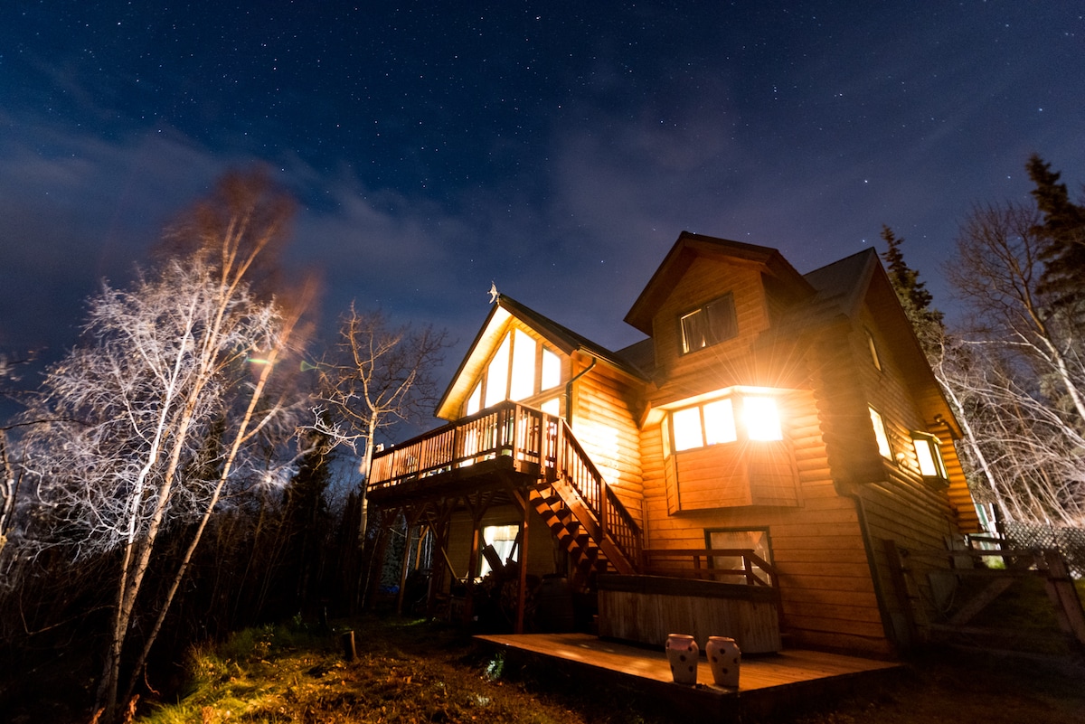 The log chalet is illuminated against a starry night sky, featuring multiple lit windows that highlight its charming architecture. The exterior includes a wooden deck with stairs leading up to the main entrance, surrounded by trees and a tranquil atmosphere.