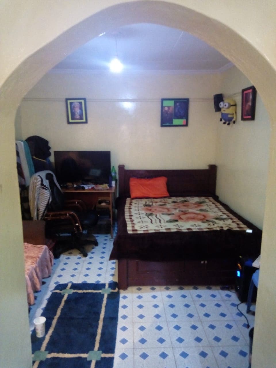 A bedsitter is visible through an arched entry. A spacious bed with a floral blanket occupies one side, while a small television is positioned on a stand nearby. A desk chair is noted alongside a computer setup. The floor features a patterned tile design and a small rug.