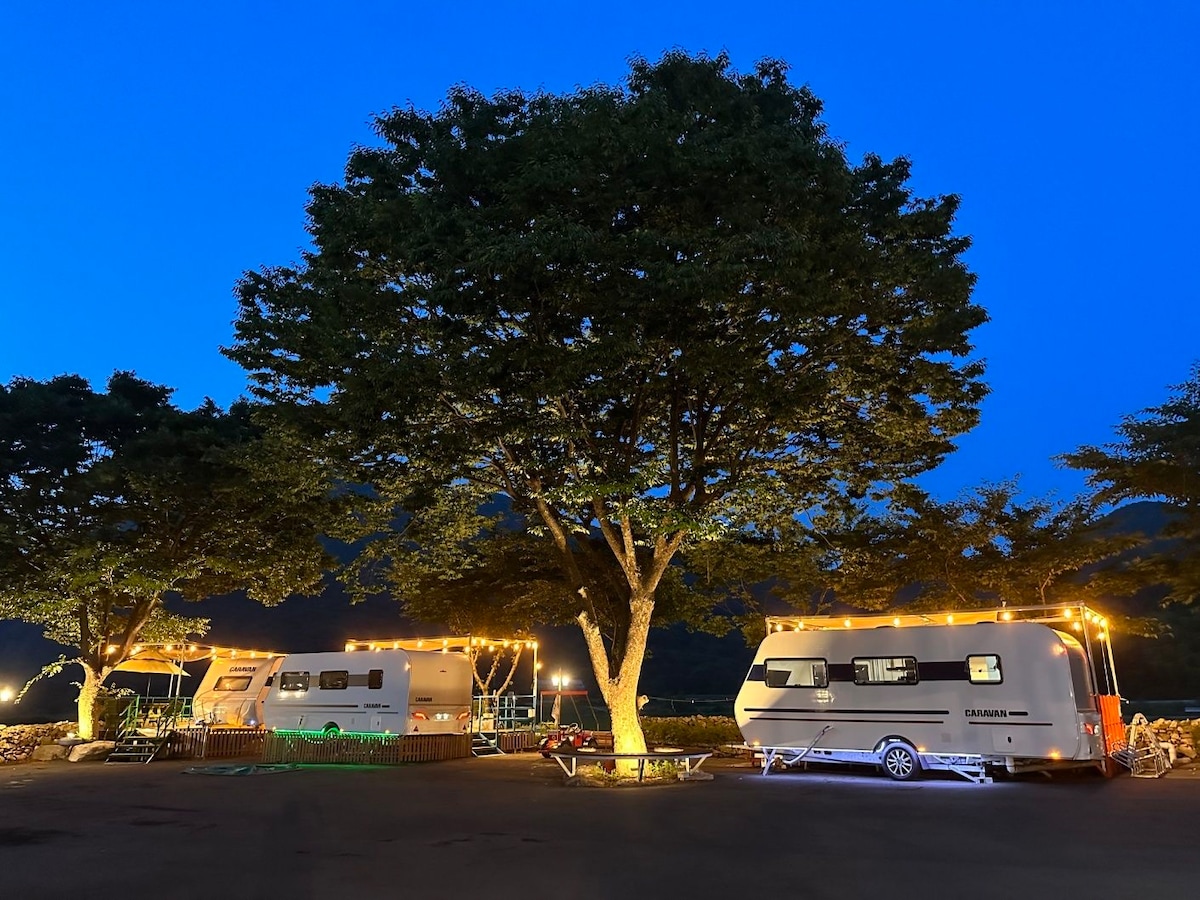 Two modern caravans are illuminated by soft lighting, nestled beneath a large tree. The scene is set at dusk, highlighting the peaceful surroundings. Outdoor seating is visible in the foreground, creating an inviting atmosphere for relaxation.