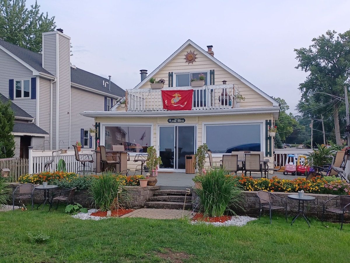 A charming lakefront house is presented with a patio adorned with colorful flowers and neatly arranged outdoor seating. Expansive windows provide views of the water, while a flag decorates the front, contributing to the welcoming appearance of the home.