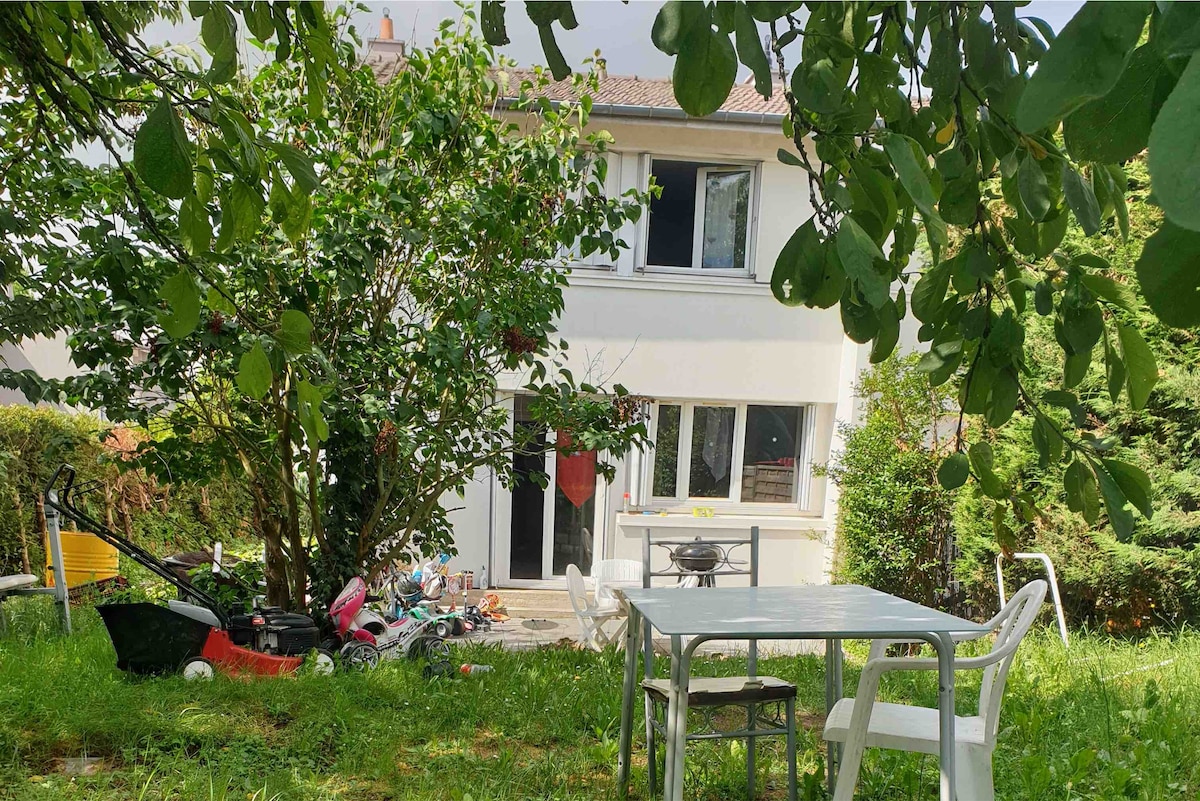The exterior of a two-story house is set against a backdrop of lush greenery. A patio area is visible, featuring a table and chairs. Nearby, a lawn mower and children's toys are scattered in the grass. Windows are seen on the upper level, allowing natural light inside.