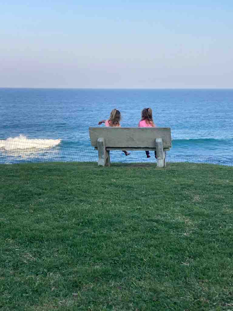Two individuals are seated on a concrete bench overlooking the ocean. The calm sea is visible in the background, with gentle waves lapping against the shore. Lush green grass surrounds the bench, providing a serene atmosphere.