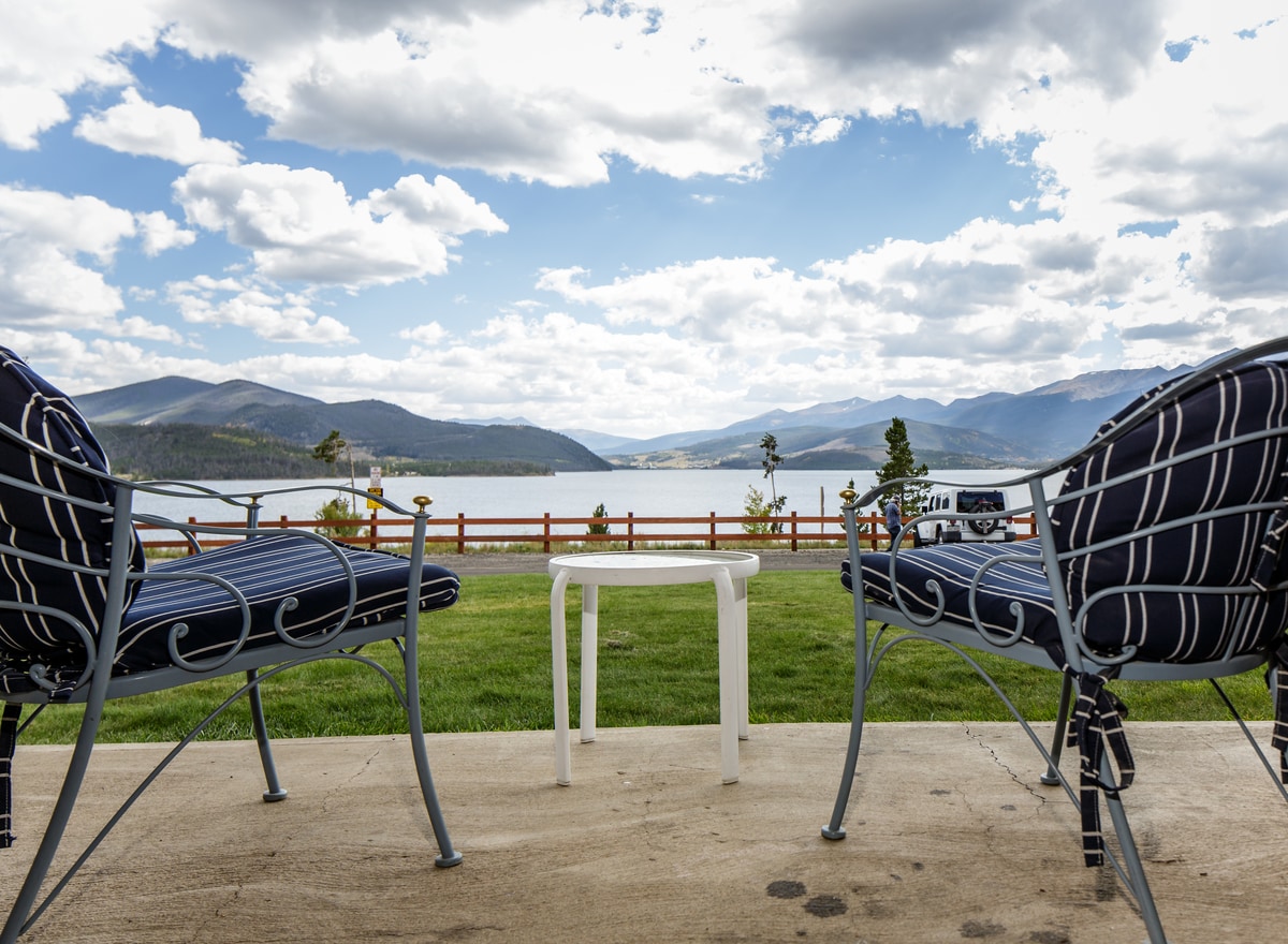 Two chairs are positioned on a concrete patio, facing a scenic view of the lake and mountains. A small white table is placed between the chairs. The backdrop features a vibrant sky filled with clouds and rolling hills in the distance.