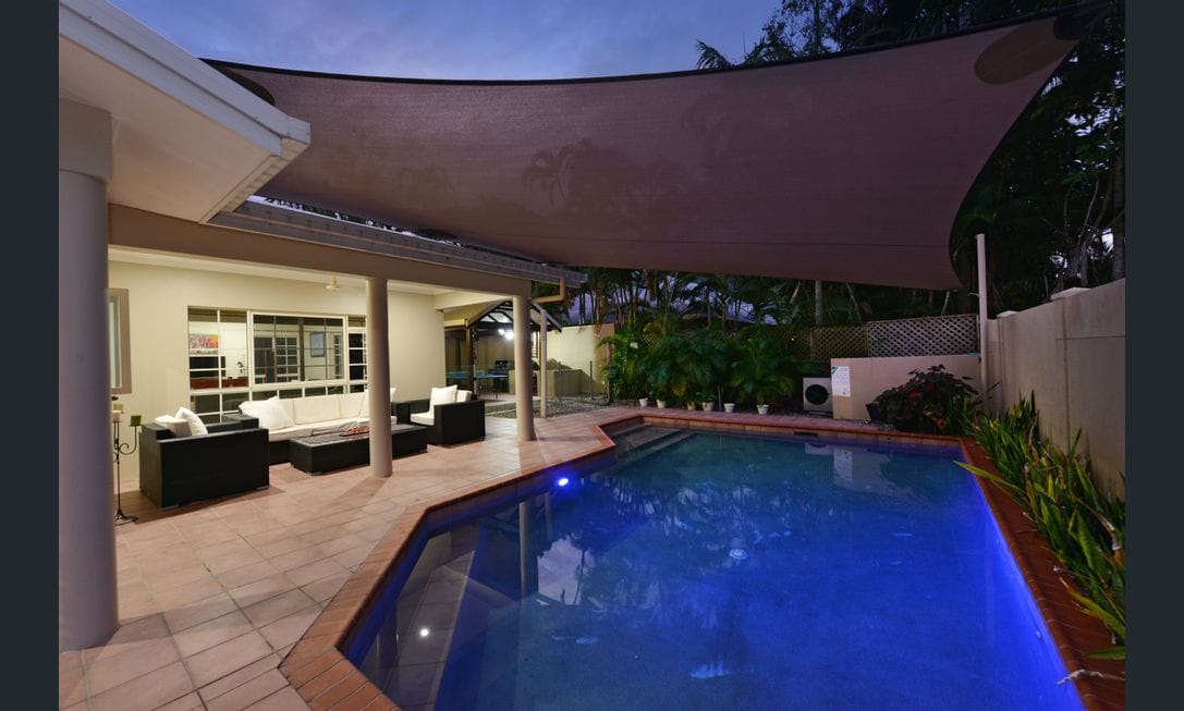 An outdoor lounge area is featured beside a swimming pool, illuminated in soft blue tones. A large shade sail provides coverage over the seating arrangement, which includes dark-colored sofas. Lush tropical greenery surrounds the space, enhancing the serene atmosphere.