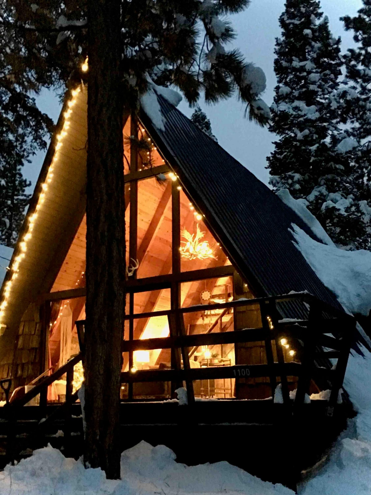 The A-frame cabin is framed by tall pine trees and glows warmly from within as evening descends. Soft lighting outlines the structure, highlighting the chalet-style architecture against a snowy backdrop. The inviting ambiance suggests a cozy retreat amidst the natural surroundings.
