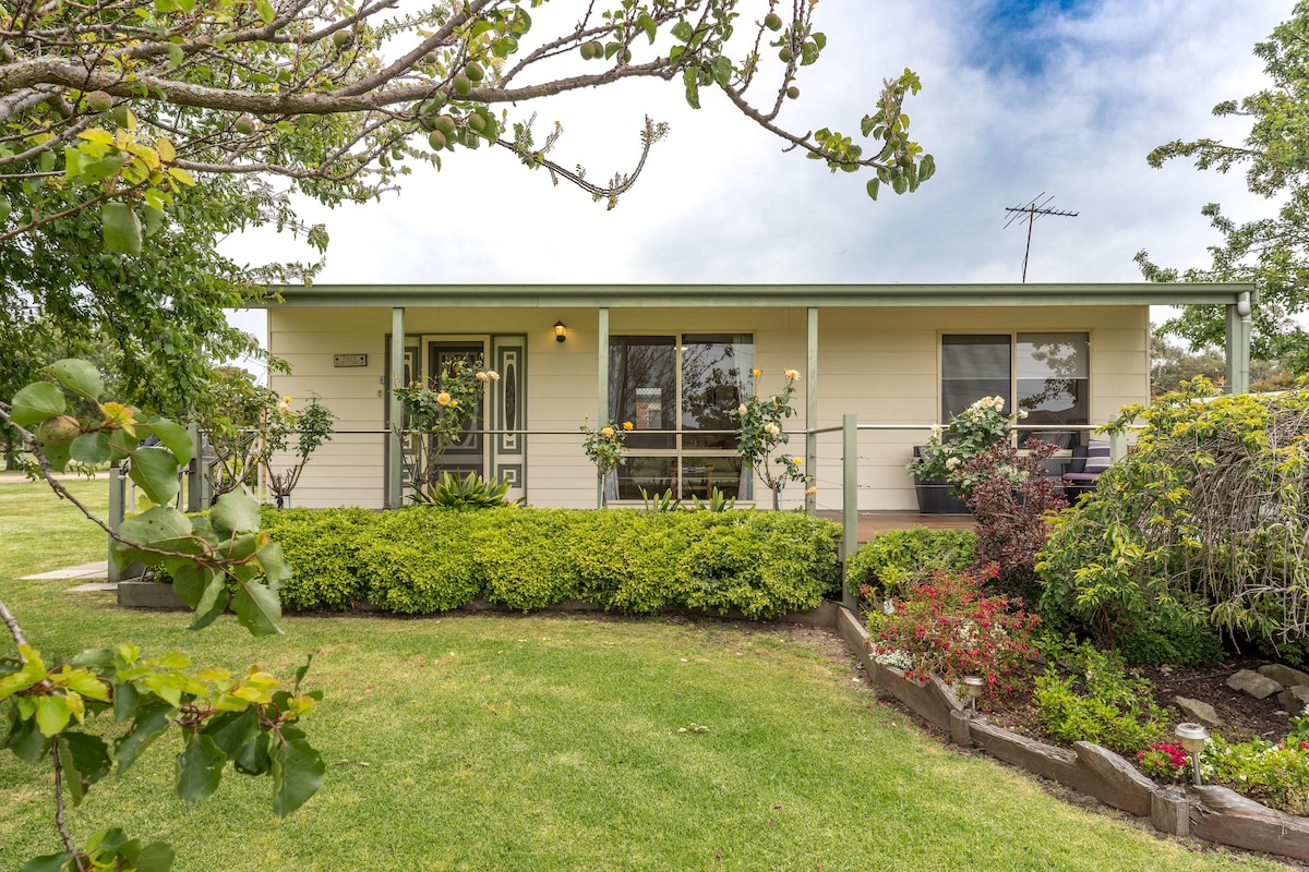 A charming cottage is presented with a welcoming front porch, surrounded by neatly trimmed hedges and flowering plants. The landscape features vibrant greenery and a variety of shrubs, with a clear sky above, enhancing the peaceful rural setting.
