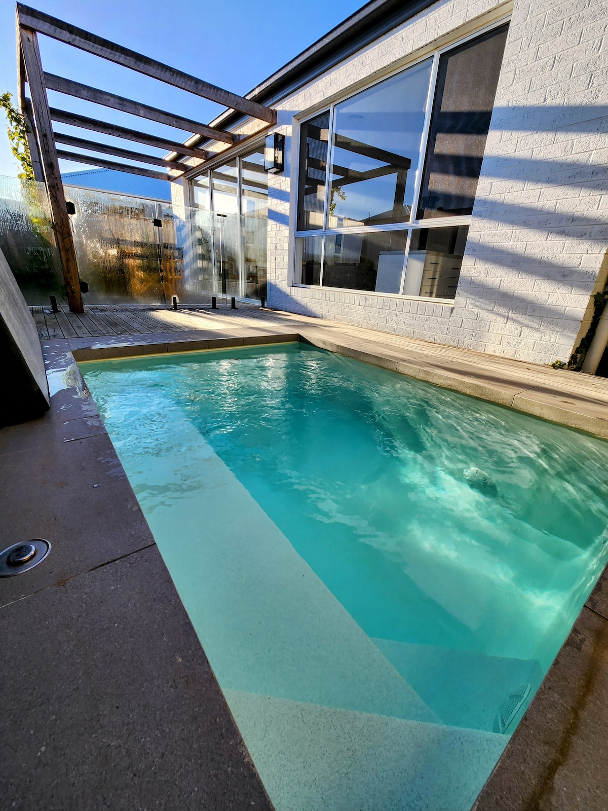 A heated plunge pool is framed by a wooden deck and large glass panels, allowing natural light to brighten the area. The pool features stairs leading in, with clear blue water reflecting the sky above.