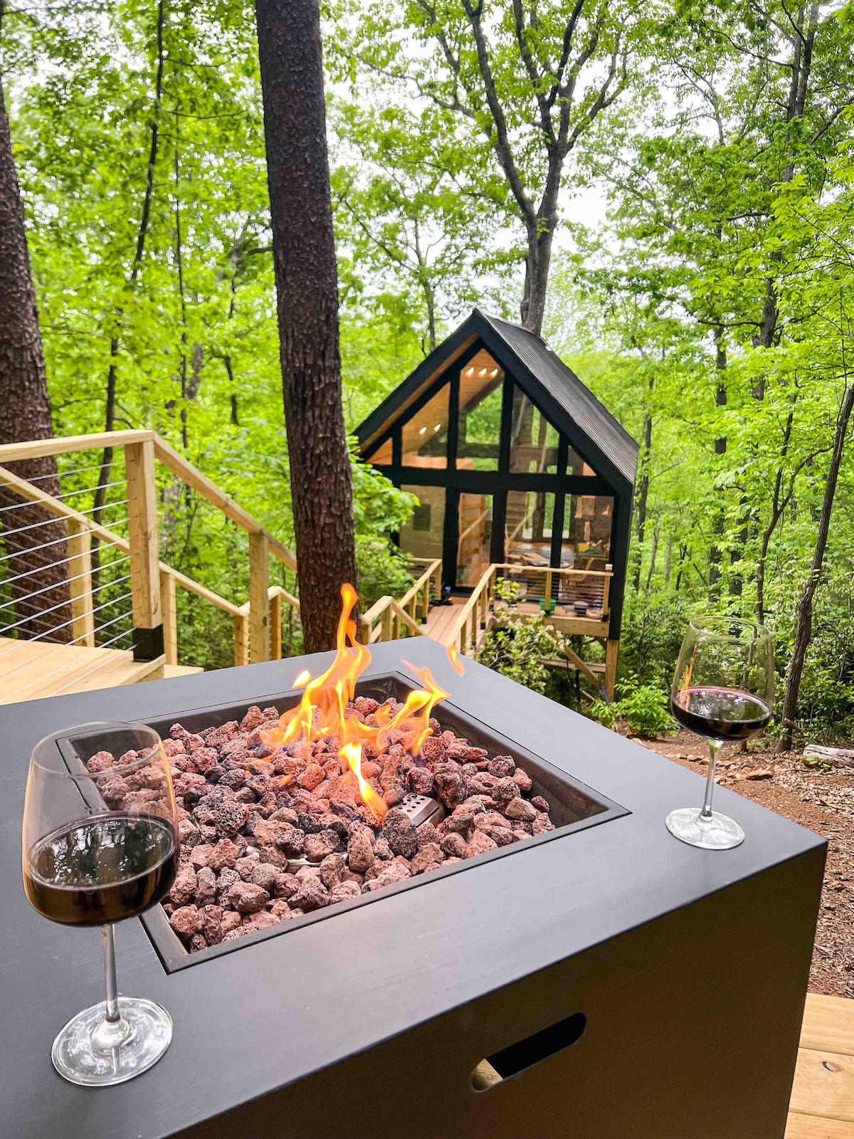 A modern fire pit sits in the foreground, surrounded by stones and flames gently flickering. Two wine glasses rest beside it, with a view of the glass-fronted treehouse in the background, framed by lush greenery and trees.