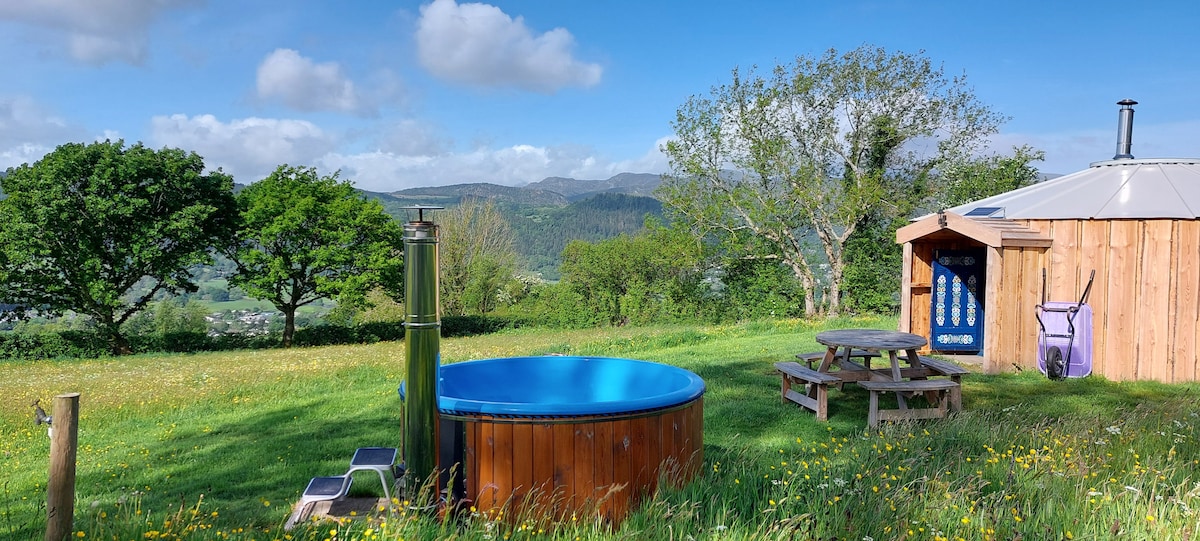 A wood-fueled hot tub is situated in a grassy area, surrounded by blooming wildflowers. A picnic table is nearby, with rolling hills and trees forming the scenic backdrop. The yurt, with its wooden cladding and chimney, is visible in the distance against a clear sky.