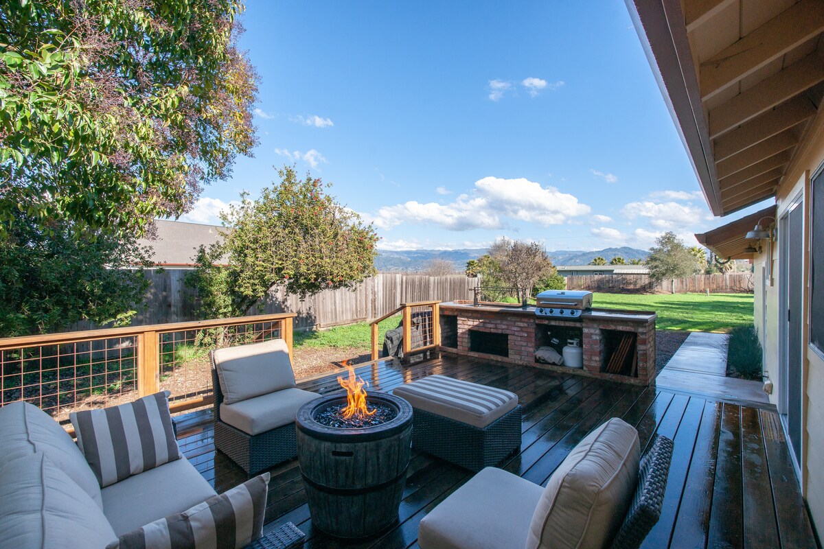 An outdoor deck features a cozy seating area with striped cushions around a fire pit. A barbeque grill is positioned nearby, while a spacious grassy lawn extends to the back. Lush greenery and distant views of hills enhance the serene setting.