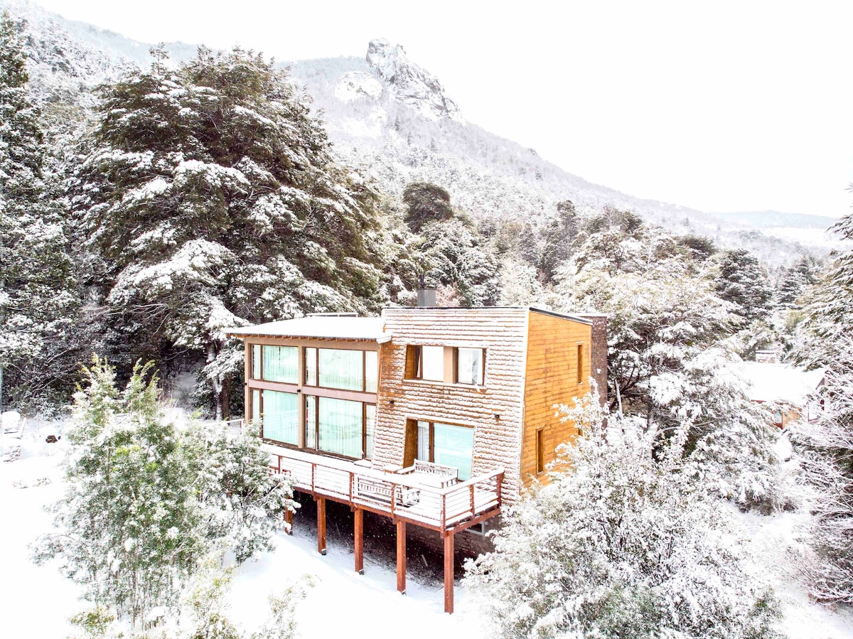 A modern mountain house is surrounded by a blanket of freshly fallen snow. Large windows invite natural light, offering views of the snowy landscape. The wooden deck extends outward, allowing for outdoor relaxation while nestled within the tranquil scenery of towering trees and distant mountains.