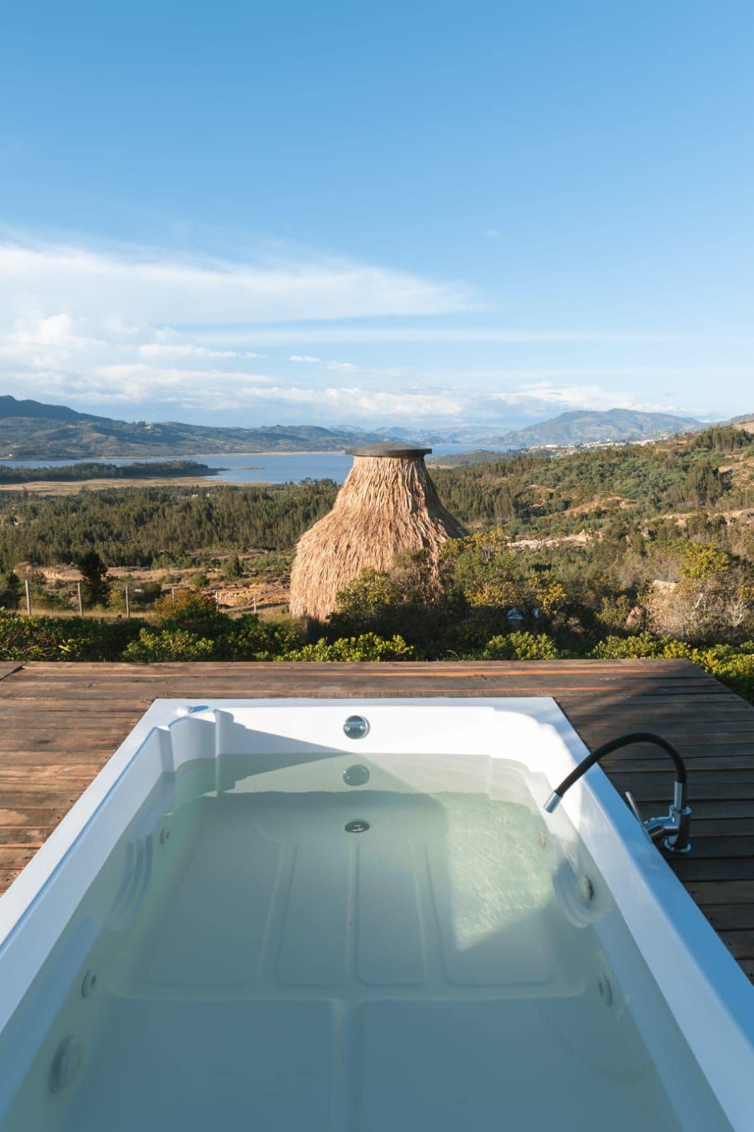 A private jacuzzi is positioned on a wooden deck, overlooking a vast landscape. In the background, a unique structure rises amidst rolling hills and a serene body of water, with a clear blue sky above. Natural greenery complements the tranquil setting.