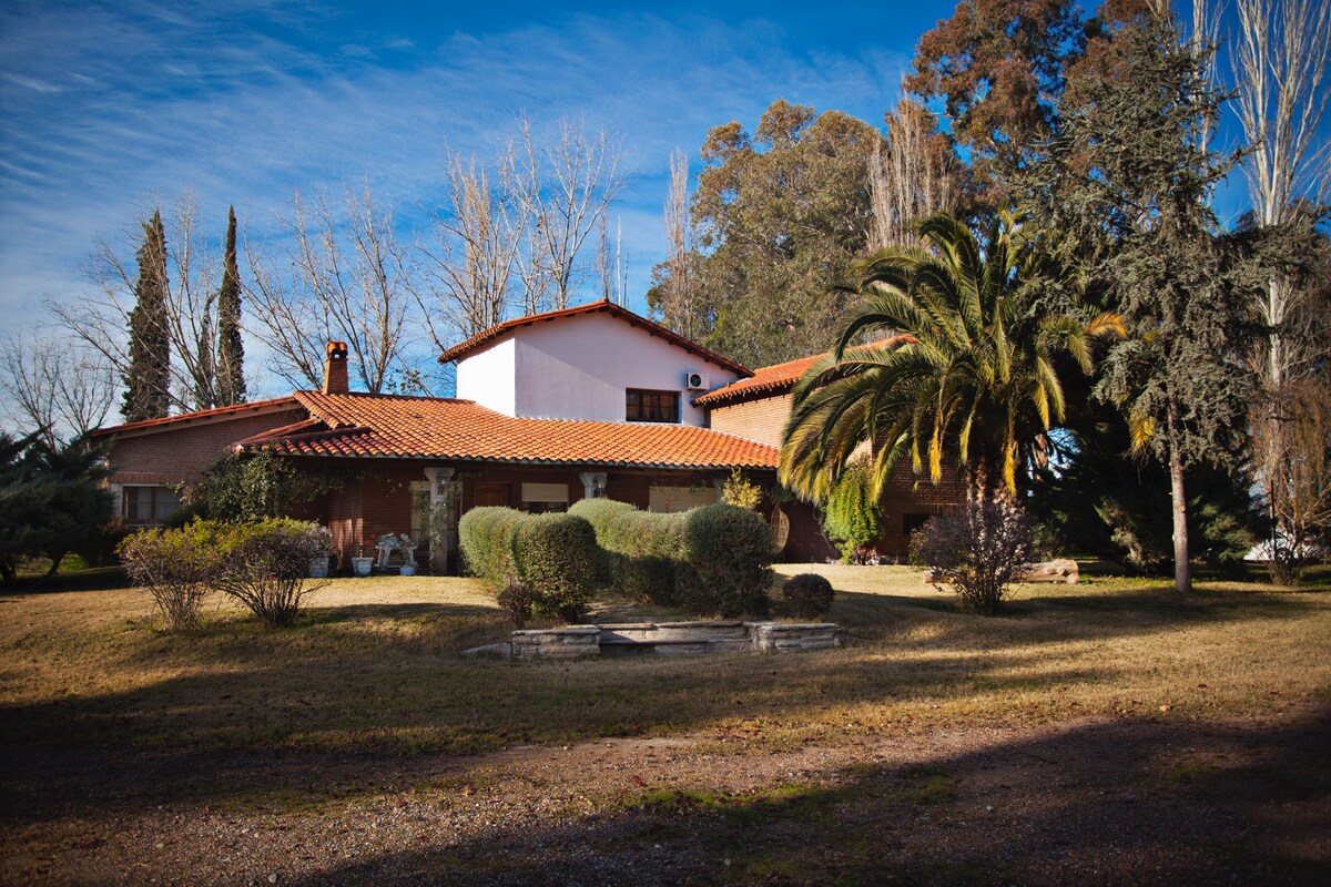 A spacious chalet is surrounded by lush greenery and mature trees. The building features a tiled roof and a mix of brick and plaster exteriors. The landscaped yard includes hedges and stone pathways, creating a welcoming outdoor space.