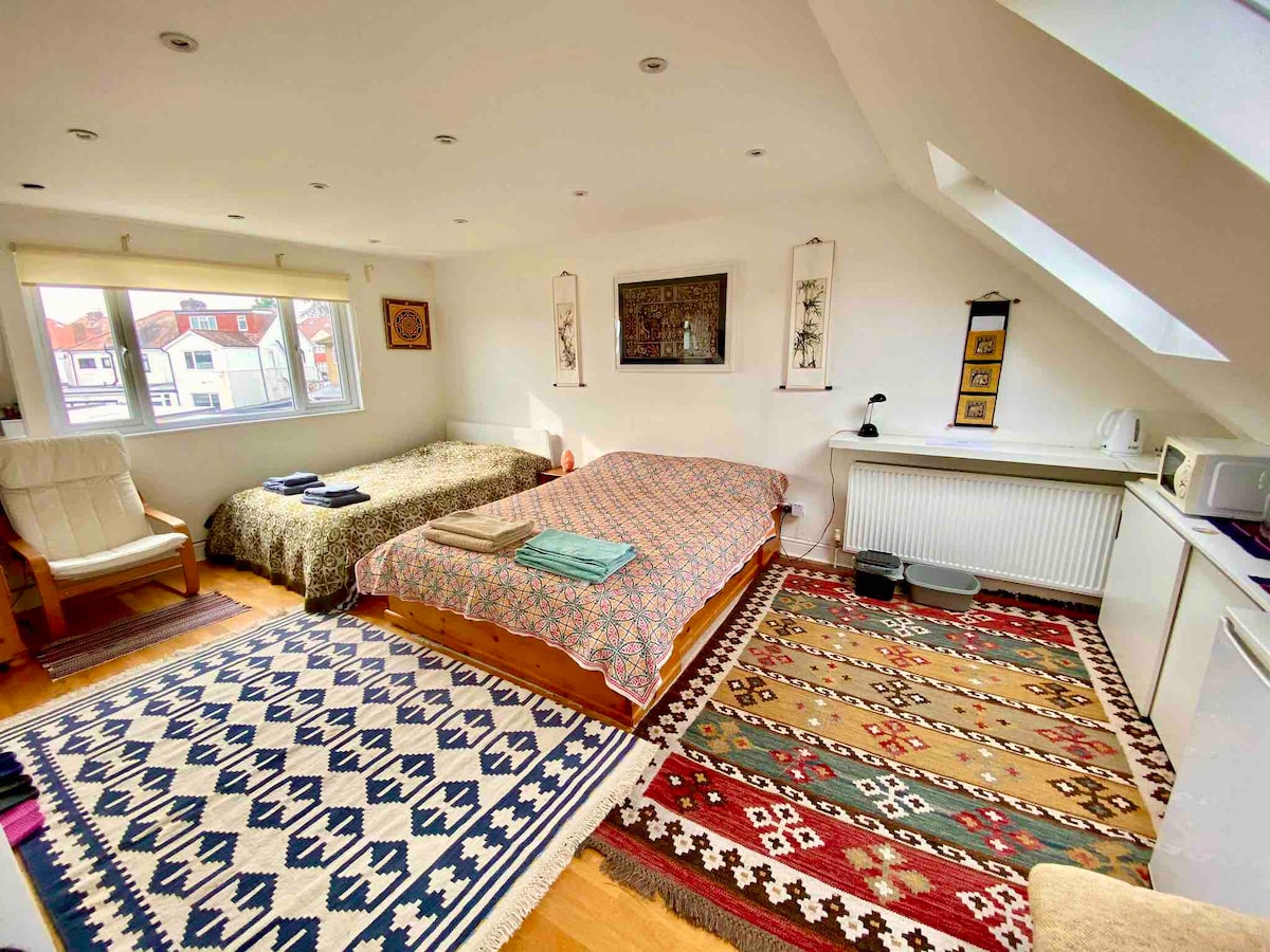 A spacious room features two double beds adorned with colorful patterned bedding. Natural light fills the space through large windows. A comfortable rocking chair is positioned beside a neatly arranged utility area with a kettle and microwave, alongside decorative rugs enhancing the room's warmth.