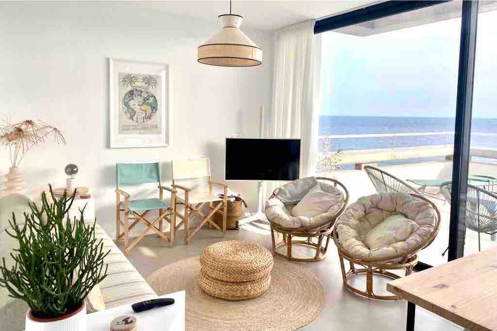 The living room features a light, airy design with large windows overlooking the ocean. A plush, circular rug is placed beneath a coffee table. Two lounge chairs and a built-in sofa provide comfortable seating, while a minimalist television is positioned nearby.