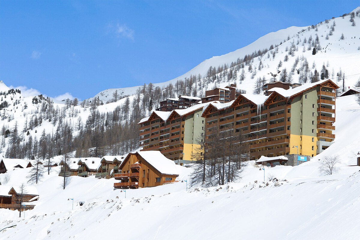 A modern apartment complex is nestled in a snowy landscape, surrounded by gently sloping mountains. The building features a combination of stone and wood accents, with balconies offering potential views of the surrounding winter scenery. The area is blanketed in fresh snow, providing a serene winter atmosphere.