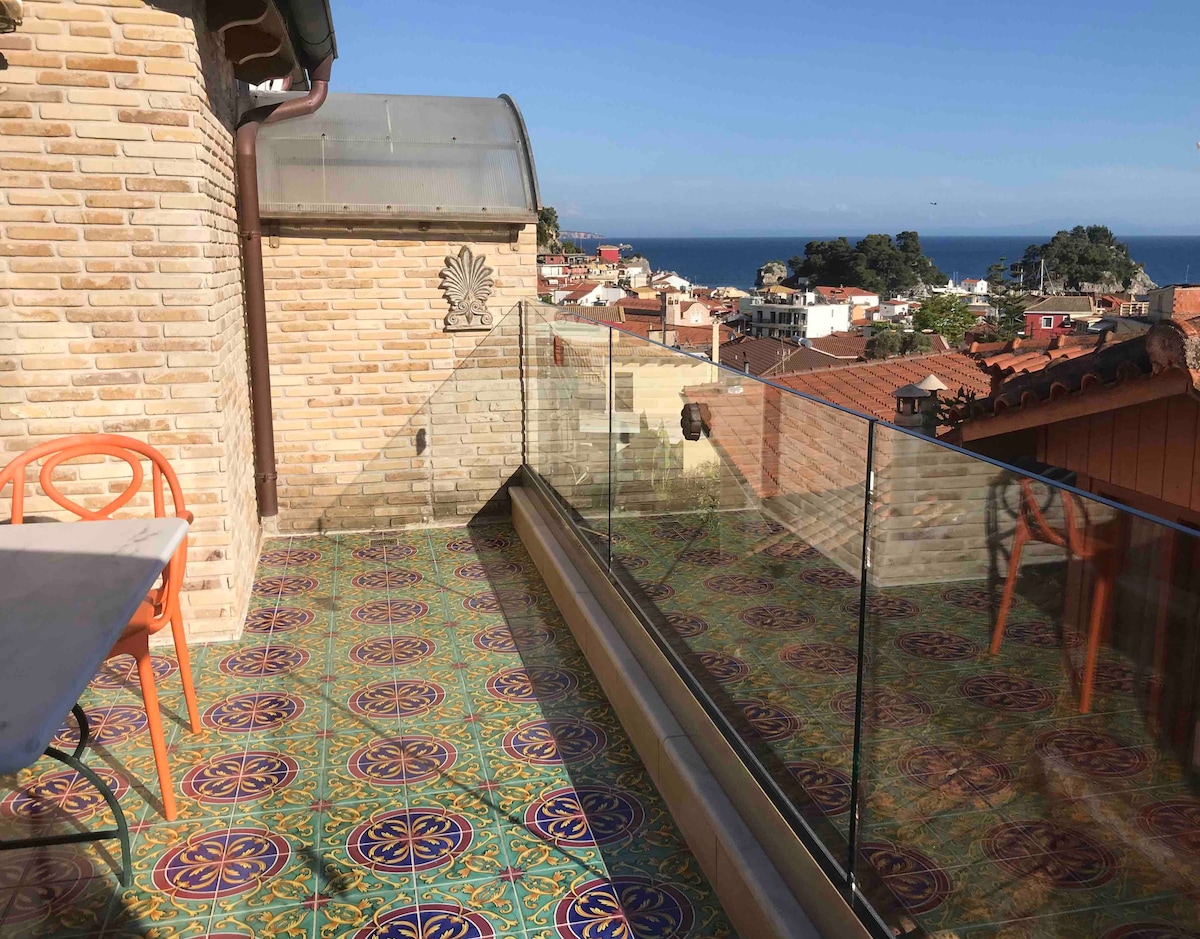 A spacious balcony features a vibrant tiled floor and glass railing, offering an unobstructed view of the sea and surrounding rooftops. The area is complemented by a small table and colorful chairs, providing a delightful setting for relaxation.